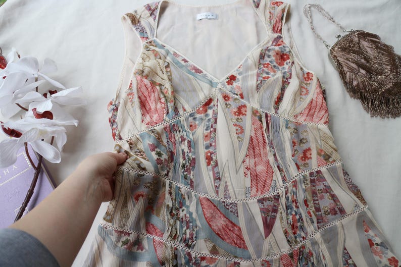 May include: A sleeveless, floral print dress with a V-neckline and tiered skirt. The dress features a mix of red, blue, and cream-colored patterns. A small, beaded purse with fringe is visible in the background.