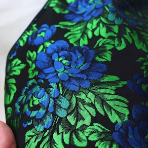 K&ouml;nnte beinhalten: Nahaufnahme eines Stoffes mit floralem Muster. Der Stoff ist schwarz mit gro&szlig;en blauen und gr&uuml;nen Blumen und Bl&auml;ttern. Die Blumen sind leuchtend blau und die Bl&auml;tter leuchtend gr&uuml;n. Der Stoff hat einen leichten Glanz.