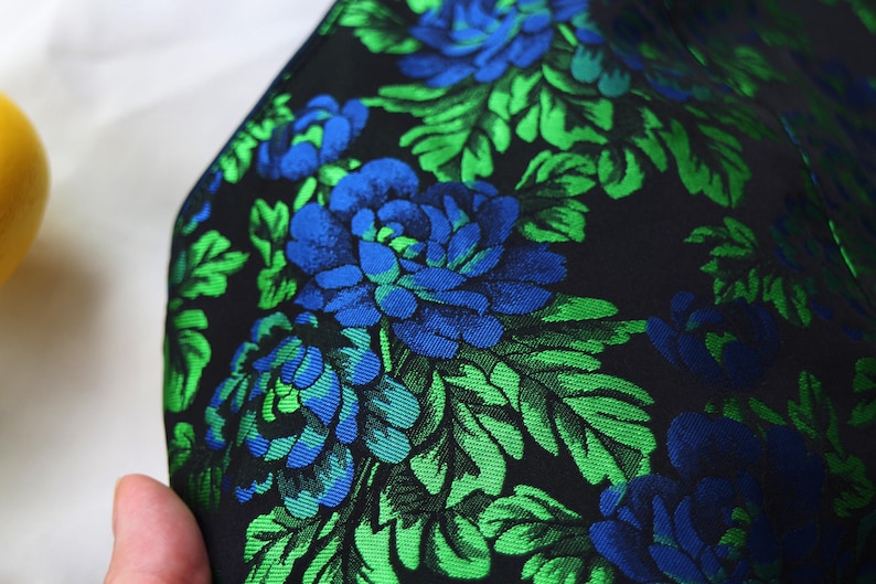 K&ouml;nnte beinhalten: Nahaufnahme eines Stoffes mit floralem Muster. Der Stoff ist schwarz mit gro&szlig;en blauen und gr&uuml;nen Blumen und Bl&auml;ttern. Die Blumen sind leuchtend blau und die Bl&auml;tter leuchtend gr&uuml;n. Der Stoff hat einen leichten Glanz.