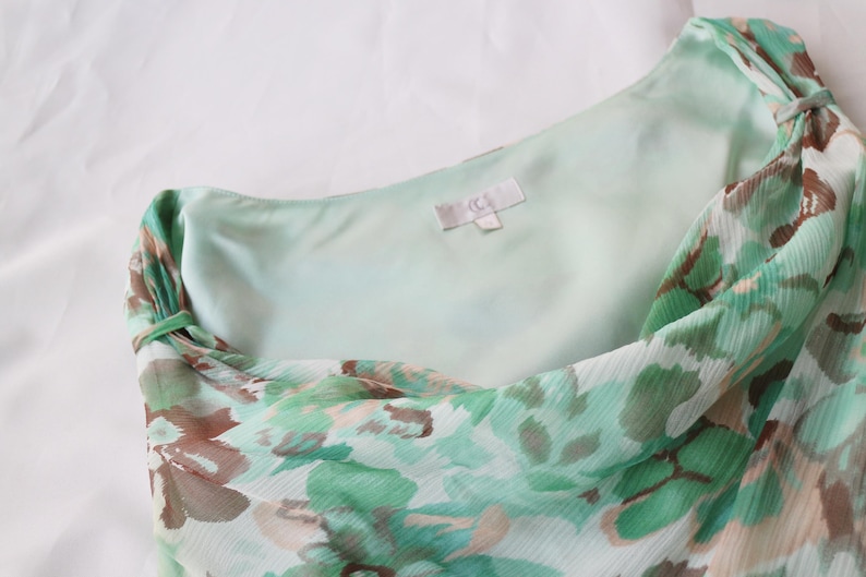 May include: A light green and brown floral print dress. The dress features a one-shoulder design with a flowing, lightweight fabric. The inner lining is a solid light green color. The dress has a tag with the letters "jc".