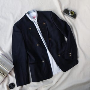 May include: A navy blue blazer with gold buttons and a white collared shirt underneath. The blazer has a double-breasted design and is laid out on a cream-colored fabric. A vintage camera is also visible.