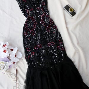 May include: Black evening gown with a V-neck and cap sleeves, embellished with silver sequins and red accents. The dress features a flowing, tiered skirt. A vintage camera and white flowers are also in the image.