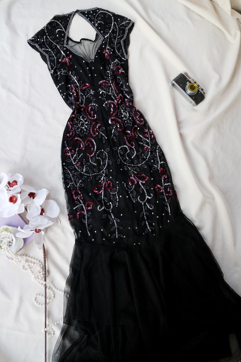 May include: Black evening gown with a V-neck and cap sleeves, embellished with silver sequins and red accents. The dress features a flowing, tiered skirt. A vintage camera and white flowers are also in the image.