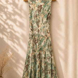 May include: A sleeveless floral print dress in shades of green, brown, and cream hangs on a wooden hanger. The dress has a flowing, flared skirt and a scoop neckline. The fabric appears lightweight and semi-sheer, with a delicate floral pattern.