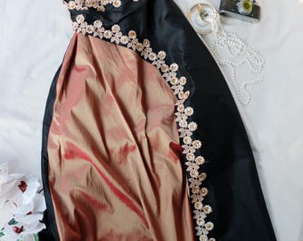 May include: A floor-length dress featuring a black bodice and a copper-toned skirt. The garment is accented with a diagonal line of cream-colored floral lace trim that extends from the bodice down the length of the skirt.