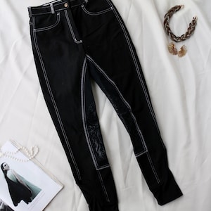 May include: Black riding pants with white stitching and decorative patches. The pants have a high waist and a classic design. A magazine, a necklace, and earrings are also in the image.