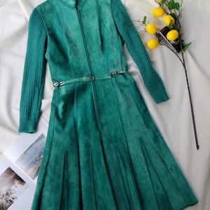 May include: A teal green suede dress with a high collar and long sleeves. The dress features a zipper closure, a belt with a silver buckle, and a pleated skirt. A sprig of lemons rests nearby.