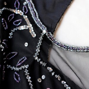 May include: Close-up of a black garment with intricate bead and sequin embellishments. Silver sequins and purple beads are arranged in floral and linear patterns. The neckline is trimmed with small purple beads.