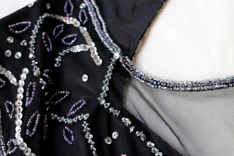 May include: Close-up of a black garment with intricate bead and sequin embellishments. Silver sequins and purple beads are arranged in floral and linear patterns. The neckline is trimmed with small purple beads.