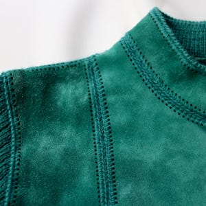 May include: Close-up of a teal jacket featuring a combination of knit and suede-like textures. The jacket has a ribbed knit sleeve and collar, with decorative stitching along the seams and a zipper closure.