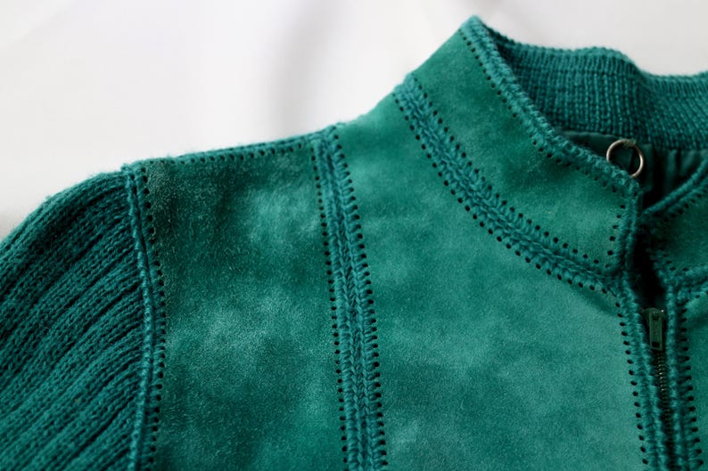 May include: Close-up of a teal jacket featuring a combination of knit and suede-like textures. The jacket has a ribbed knit sleeve and collar, with decorative stitching along the seams and a zipper closure.