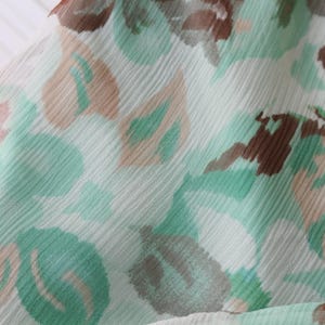 May include: Close-up of a fabric with a floral pattern. The design features soft green, brown, and beige shapes on a white background. The fabric has a slightly textured appearance, suggesting a lightweight material.