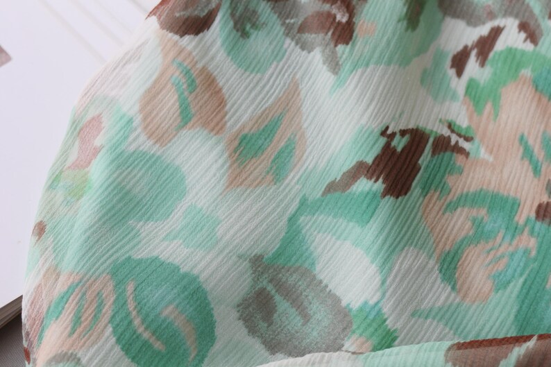 May include: Close-up of a fabric with a floral pattern. The design features soft green, brown, and beige shapes on a white background. The fabric has a slightly textured appearance, suggesting a lightweight material.