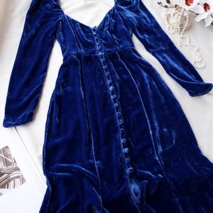 May include: A long, royal blue velvet dress with long sleeves and a button-down front. The dress features a sweetheart neckline and a flowing skirt. The fabric has a soft, luxurious texture. The dress is laid out on a white surface.