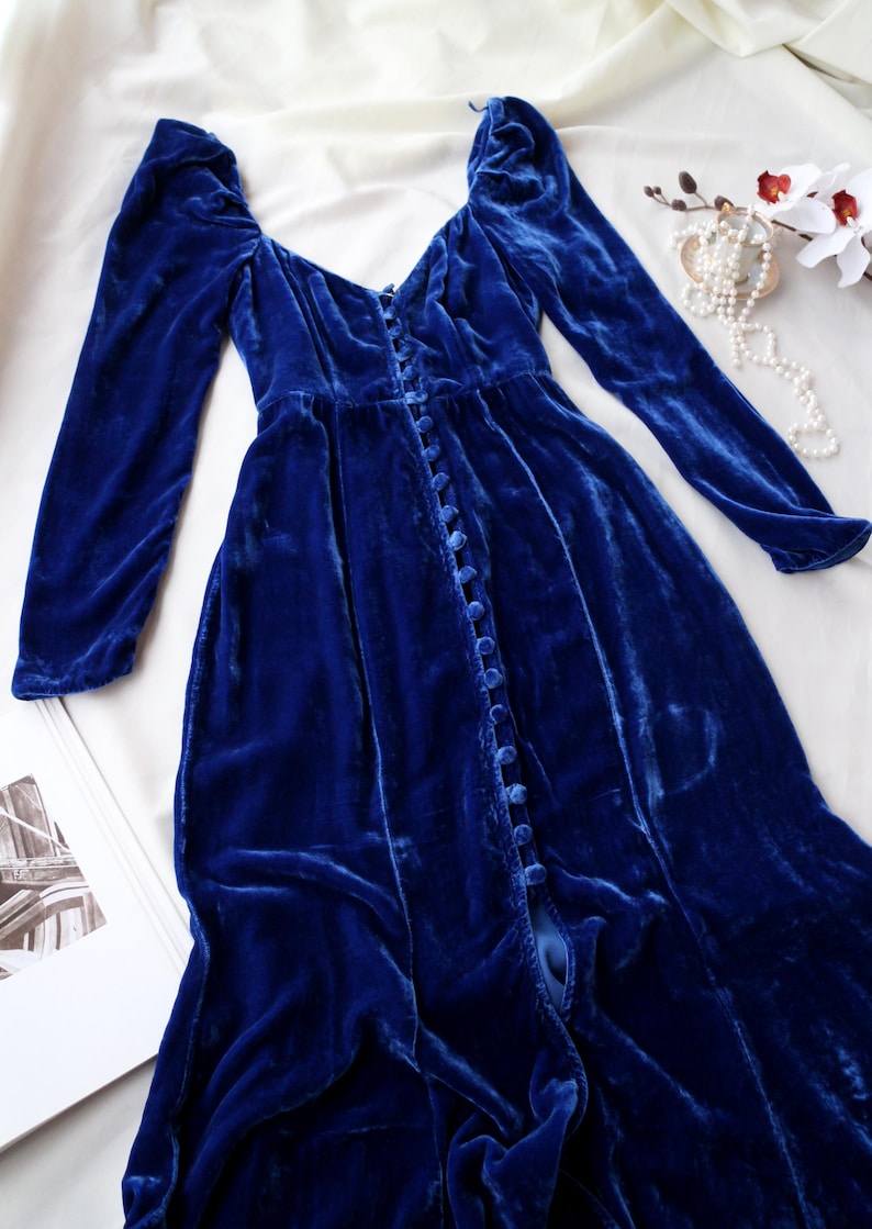May include: A long, royal blue velvet dress with long sleeves and a button-down front. The dress features a sweetheart neckline and a flowing skirt. The fabric has a soft, luxurious texture. The dress is laid out on a white surface.