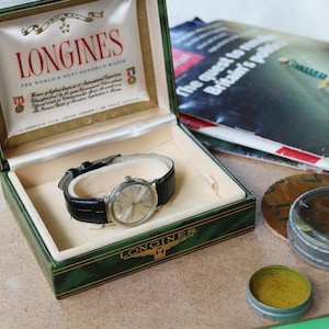 May include: A silver-toned Longines wristwatch with a black leather strap rests inside a green hinged presentation box. The box interior features white satin lining with the text Longines The World's Most Honored Watch and various award icons.