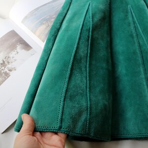 May include: Close-up of a teal green velvet skirt with a pleated design. The skirt has vertical seams and a soft, luxurious texture. The fabric is a rich, solid color, and the stitching is visible.