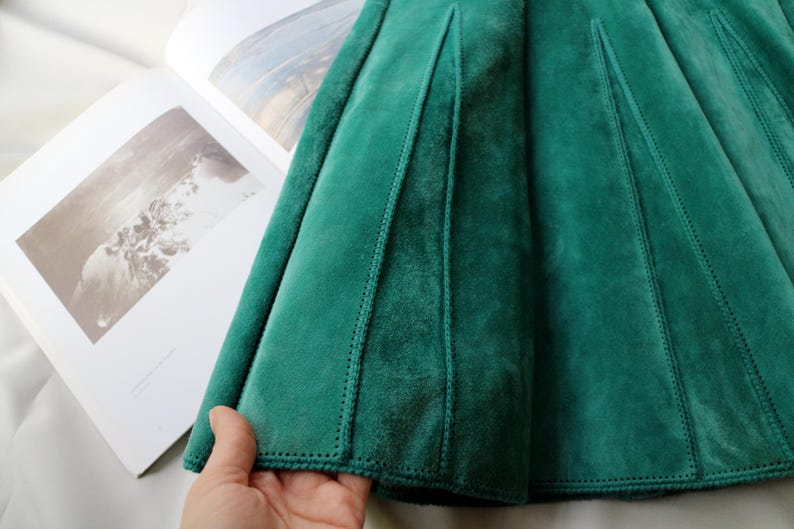 May include: Close-up of a teal green velvet skirt with a pleated design. The skirt has vertical seams and a soft, luxurious texture. The fabric is a rich, solid color, and the stitching is visible.