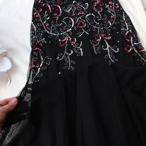 May include: Black evening gown with floral embellishments. The bodice features intricate beadwork and sequin detailing in silver and red. The skirt is made of a flowing black fabric.