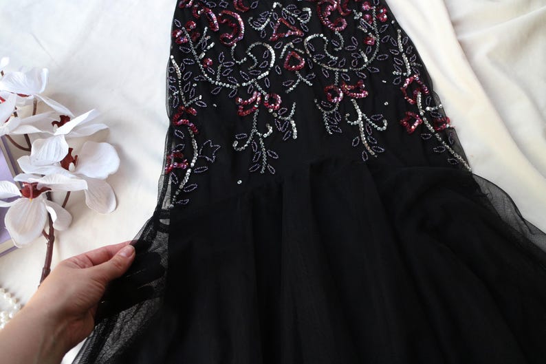 May include: Black evening gown with floral embellishments. The bodice features intricate beadwork and sequin detailing in silver and red. The skirt is made of a flowing black fabric.