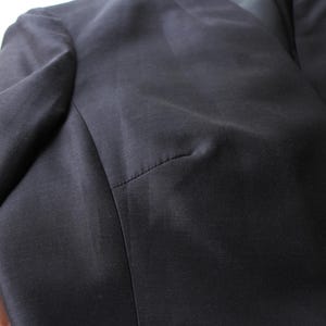 May include: Close-up of a dark navy blazer. The fabric has a subtle sheen, and the stitching is visible along the seams. A single, dark button is visible on the front. The blazer's shoulder and sleeve are in focus.
