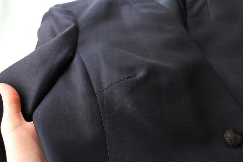 May include: Close-up of a dark navy blazer. The fabric has a subtle sheen, and the stitching is visible along the seams. A single, dark button is visible on the front. The blazer's shoulder and sleeve are in focus.