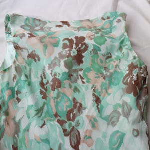 May include: A sleeveless top with a floral pattern in shades of green, brown, and beige. The fabric appears to be lightweight and semi-sheer, with a soft, flowing drape. The top is likely part of a summer clothing collection.