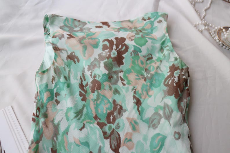 May include: A sleeveless top with a floral pattern in shades of green, brown, and beige. The fabric appears to be lightweight and semi-sheer, with a soft, flowing drape. The top is likely part of a summer clothing collection.