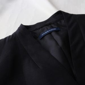 May include: Close-up of a black blazer with a notched collar. The blazer has a tailored fit and a label that reads "WINDS&MOOR". The fabric appears to be a smooth, woven material. The background is a white, textured surface.