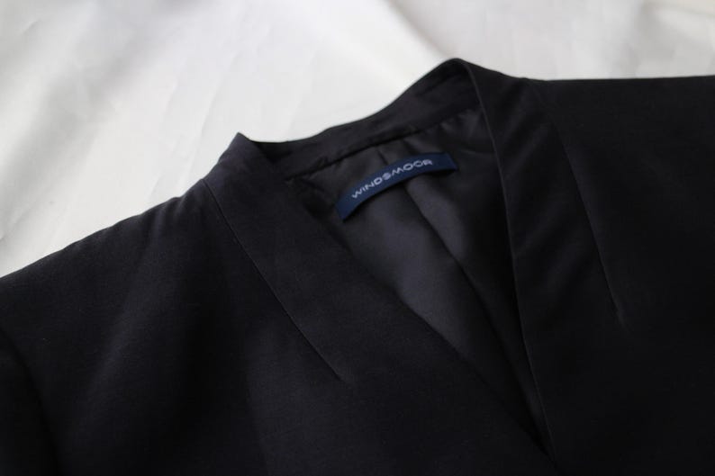 May include: Close-up of a black blazer with a notched collar. The blazer has a tailored fit and a label that reads "WINDS&MOOR". The fabric appears to be a smooth, woven material. The background is a white, textured surface.