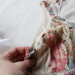 May include: A close-up of a floral patterned dress with a V-neckline. The dress features a mix of red, blue, and beige colors. The fabric appears to be lightweight and semi-sheer, with a delicate lace trim along the neckline.