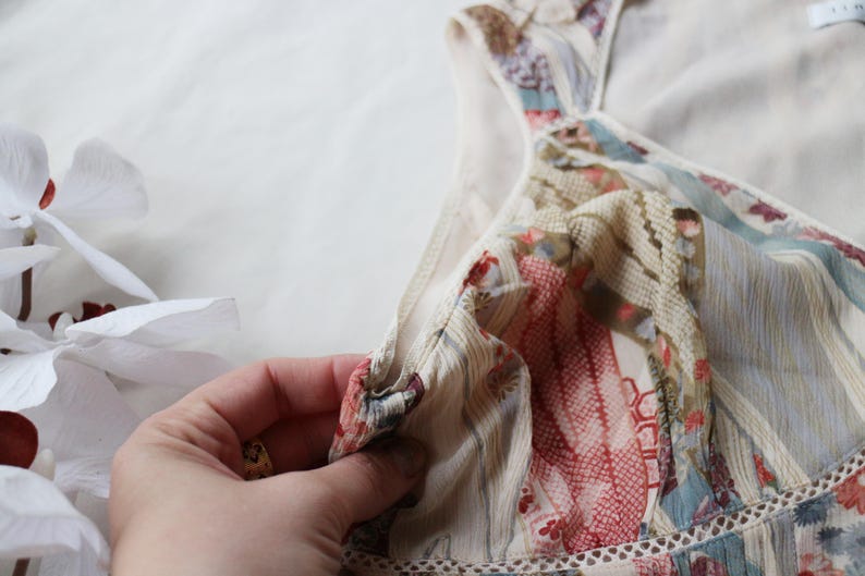 May include: A close-up of a floral patterned dress with a V-neckline. The dress features a mix of red, blue, and beige colors. The fabric appears to be lightweight and semi-sheer, with a delicate lace trim along the neckline.