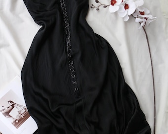 May include: Black halter dress with a beaded embellishment down the front. The dress is made of a flowing fabric and has a tie at the neck. A sprig of white orchids with red centers is next to the dress.