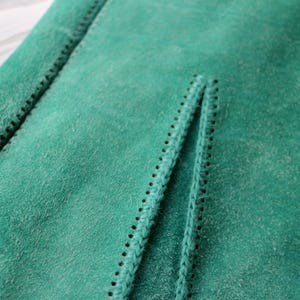 May include: Close-up of a section of green suede fabric. The fabric has a soft, textured surface with visible stitching along the edges and a central seam. The stitching is black and creates a decorative border.
