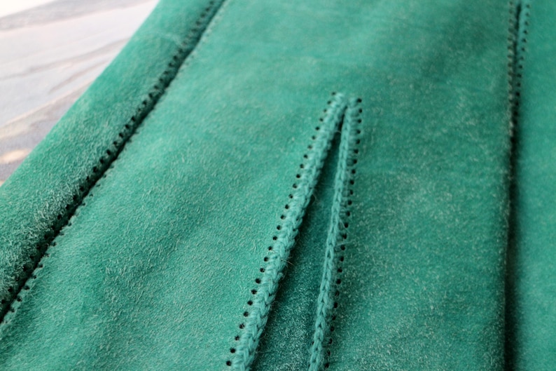 May include: Close-up of a section of green suede fabric. The fabric has a soft, textured surface with visible stitching along the edges and a central seam. The stitching is black and creates a decorative border.