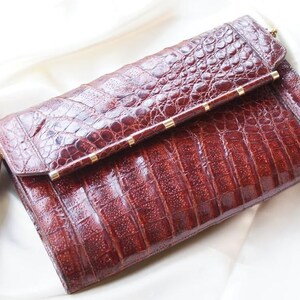 May include: A brown, textured clutch with a crocodile skin pattern. The rectangular bag has a fold-over flap secured with a gold-toned clasp and accents. The bag is resting on a white surface.