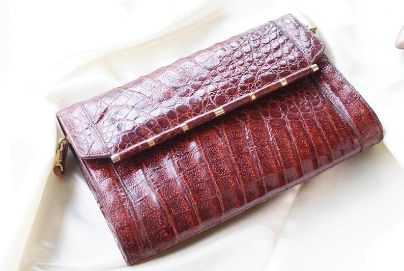 May include: A brown, textured clutch with a crocodile skin pattern. The rectangular bag has a fold-over flap secured with a gold-toned clasp and accents. The bag is resting on a white surface.