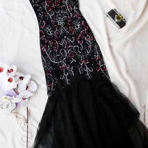 May include: A black evening gown with intricate sequin and bead embellishments in silver and burgundy. The dress features a sheer neckline and a flowing, layered skirt. A vintage camera and white flowers are also in the image.