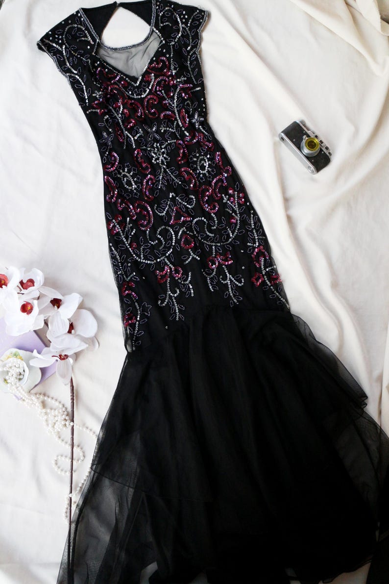 May include: A black evening gown with intricate sequin and bead embellishments in silver and burgundy. The dress features a sheer neckline and a flowing, layered skirt. A vintage camera and white flowers are also in the image.