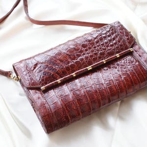 May include: A rectangular, brown leather crossbody bag with a crocodile skin texture. The bag has a flap closure with a gold-colored bar and small gold accents. A thin, brown strap is attached.