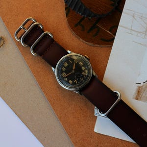 May include: A vintage wristwatch with a dark brown leather strap and a black watch face. The watch has silver-colored hardware and is set against a backdrop of tan and brown paper. The watch face displays the numbers 1 through 12.