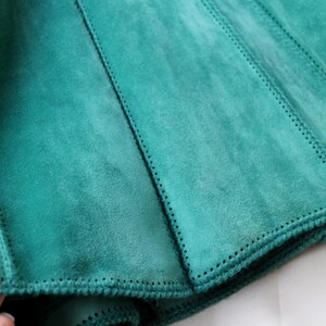 May include: Close-up of a teal suede garment, possibly a skirt or part of a jacket. The fabric is divided into panels with visible stitching. The edges are finished with a detailed, stitched border. The suede has a soft, textured appearance.