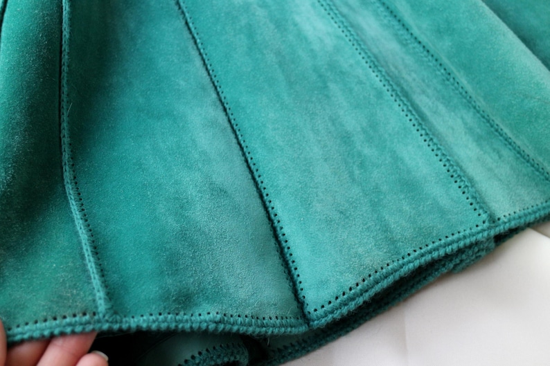 May include: Close-up of a teal suede garment, possibly a skirt or part of a jacket. The fabric is divided into panels with visible stitching. The edges are finished with a detailed, stitched border. The suede has a soft, textured appearance.