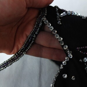 May include: Close-up of a black garment with intricate embellishments. The neckline is trimmed with small, dark beads, and the garment is adorned with sparkling sequins and beads. The fabric appears to be a sheer mesh.