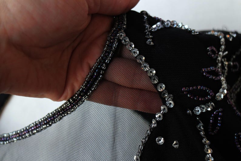 May include: Close-up of a black garment with intricate embellishments. The neckline is trimmed with small, dark beads, and the garment is adorned with sparkling sequins and beads. The fabric appears to be a sheer mesh.