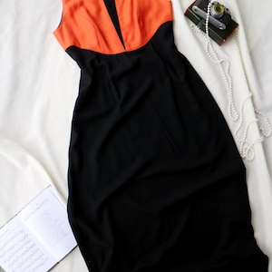May include: Sleeveless black dress with an orange bodice. The dress has a square neckline and a fitted silhouette. A vintage camera and a string of pearls are next to the dress on a white surface.