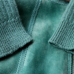 May include: Close-up of a teal-colored jacket with knitted sleeves. The jacket has a suede-like texture with visible stitching details. The sleeves are ribbed, and the zipper is visible. The overall aesthetic is cozy and textured.
