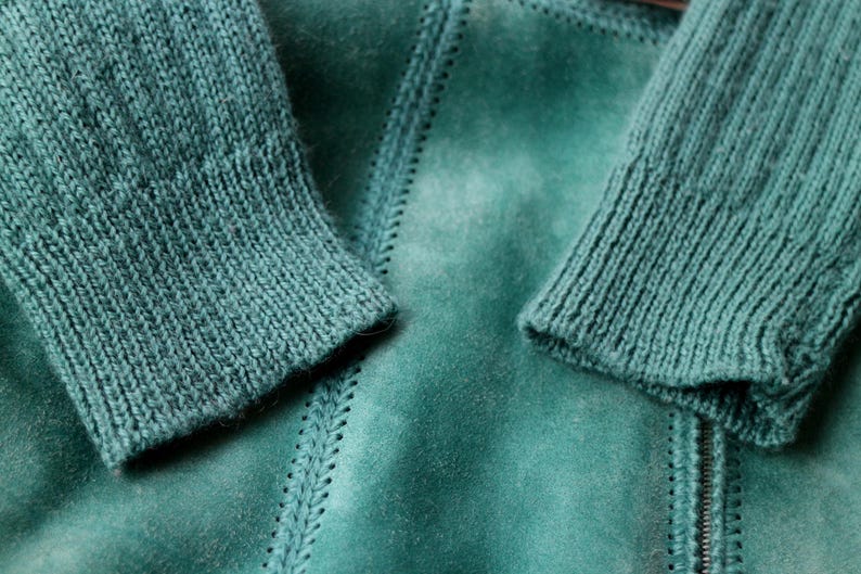 May include: Close-up of a teal-colored jacket with knitted sleeves. The jacket has a suede-like texture with visible stitching details. The sleeves are ribbed, and the zipper is visible. The overall aesthetic is cozy and textured.