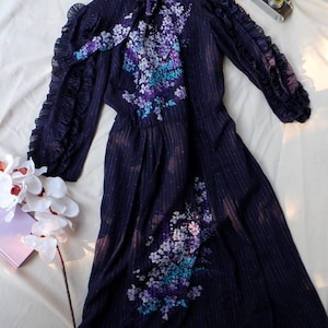 May include: A dark navy blue dress with a floral pattern in shades of purple, blue, and white. The dress features long sleeves with ruffled cuffs, a high collar, and a matching tie. A vintage camera sits nearby.