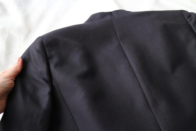 May include: Close-up of a navy blue blazer's back, showcasing the shoulder and upper back details. The fabric appears smooth and has a slight sheen. The image highlights the tailored construction of the garment, with visible seams and a classic collar.